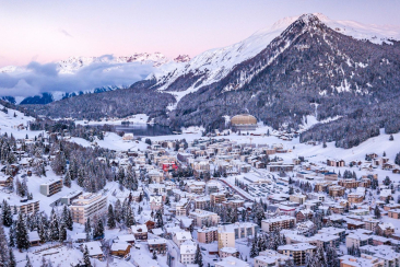 Winterpanorama von Davos mit schneebedeckten Dächern, dem AlpenGold Davos am bewaldeten Hang und den umliegenden Bündner Bergen.