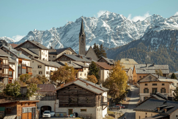 Traditionelles Engadiner Bergdorf mit Kirchturm, historischen Häusern und goldenen Lärchen vor der imposanten Kulisse schneebedeckter Gipfel.