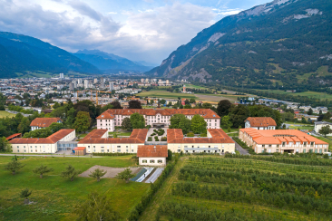 Klinikareal der Psychiatrischen Dienste Graubünden mit mehreren Gebäuden und roten Dächern in grüner Landschaft, Stadt und schneebedeckte Berge im Hintergrund.