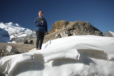 Eine Forscherin steht mit einem Laptop auf einem Gletscher, umgeben von schneebedeckten Gipfeln und Felslandschaften unter tiefblauem Himmel.