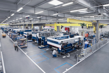 Weitläufige industrielle Produktionshalle mit automatisierten Anlagen, Laufkränen und Fachkräften bei der Montage komplexer Maschinen.