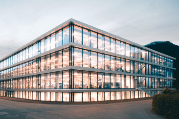 Modernes, mehrstöckiges Firmengebäude von Hamilton mit vollständig verglaster Fassade und beleuchteten Arbeitsplätzen vor dunkler Bergkulisse.