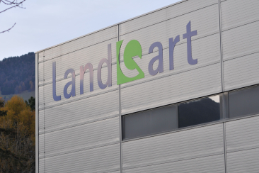 Grosses LandQart-Logo an der hellgrauen Metallfassade eines Firmengebäudes vor einer herbstlich bewaldeten Hügellandschaft.