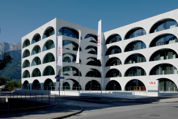 Modernes ÖKK-Gebäude mit weisser Fassade, markanten Rundbogenfenstern und Fahnen vor einer sommerlichen Bündner Berglandschaft.