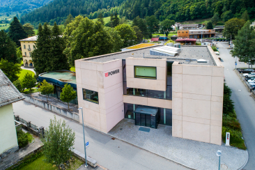 Repower-Gebäude mit grauem Betonbau und großem Logo in alpinem Tal, umgeben von dichten Fichtenwäldern, Fluss und Berglandschaft im Hintergrund.