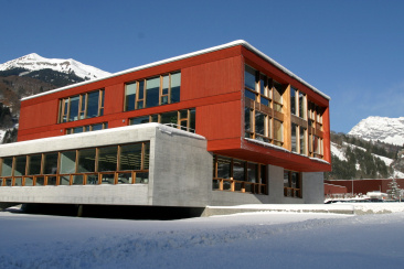 Trumpf: Modernes Gebäude mit roter Metallfassade und Betonuntergeschoss in verschneiter Alpengegend, schneebedeckte Berge im Hintergrund.