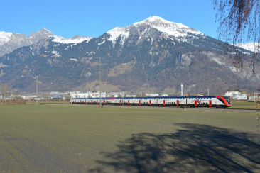 Ein SBB-Zug fährt durch die offene Landschaft bei Landquart, vor dem Panorama der verschneiten Bündner Berge und bewaldeten Hänge.