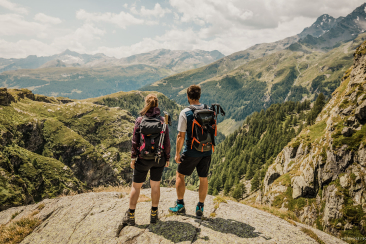 Zwei Bergsteiger stehen mit Rucksäcken auf einem Gipfel und blicken über ein weites alpines Bergpanorama mit Felsen und Wäldern.