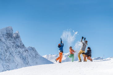 Eine Familie wirft auf einer verschneiten Piste jubelnd Schnee in die Luft, umgeben von eindrucksvollen Gipfeln unter blauem Winterhimmel.