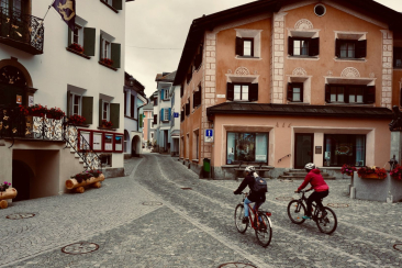 Zwei Velofahrende fahren durch die gepflasterte Gasse eines Engadiner Dorfs in der Region Maloja, vorbei an Häusern mit Sgraffiti-Fassaden.