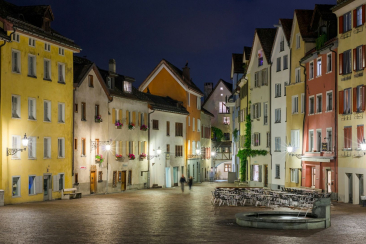 Nächtlicher Blick über einen Platz in der Churer Altstadt mit beleuchteten, farbigen Häuserfassaden, Brunnen und leeren Restaurantterrassen.