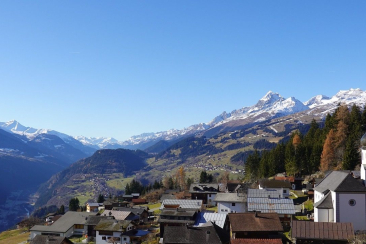 Weitblick über die Surselva mit einem Bergdorf und seiner Kirche im Vordergrund, bewaldeten Hängen sowie schneebedeckten Gipfeln am Horizont.