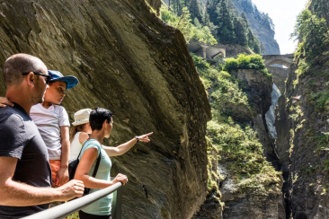 Eine Familie bestaunt von einem Steg aus die tief eingeschnittene Viamala-Schlucht mit steilen Felswänden, Wasserfall und historischen Brücken.