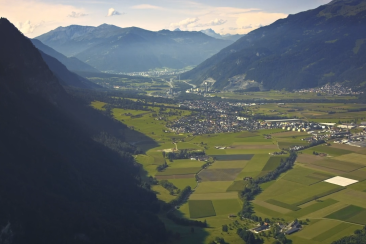 Panorama des Bündner Rheintals mit Feldern, einem Dorf und bewaldeten Berghängen vor der Kulisse der Alpen.